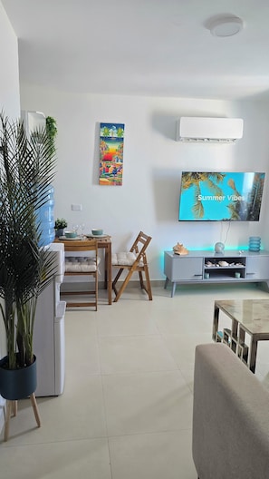Een woonkamer met een flatscreen televisie, een eethoek met een houten tafel en stoelen, en een grote kamerplant.