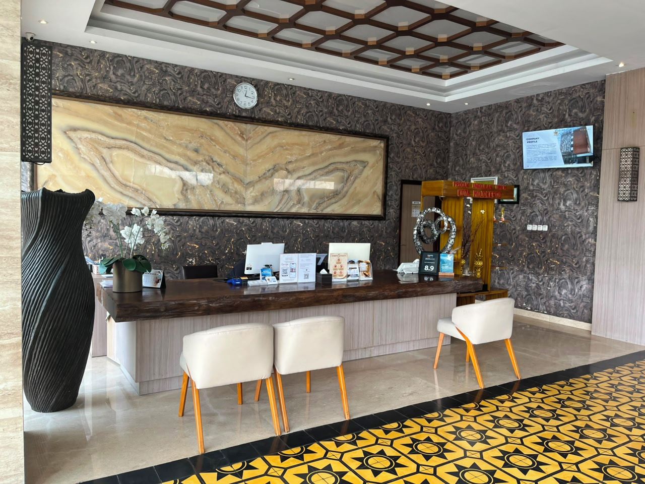 A hotel reception area with a marble wall, a clock, a reception desk, and a seating area with chairs.