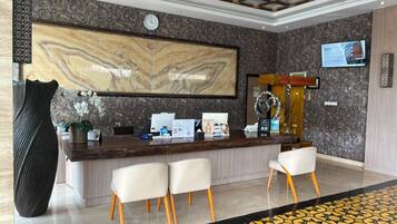 A hotel reception area with a marble wall, a clock, a reception desk, and a seating area with chairs.
