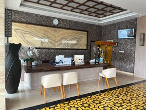 A hotel reception area with a marble wall, a clock, a reception desk, and a seating area with chairs.
