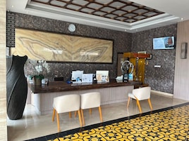 Zona de recepción de un hotel con pared de mármol, un reloj, mostrador de recepción y un área de estar con sillones.