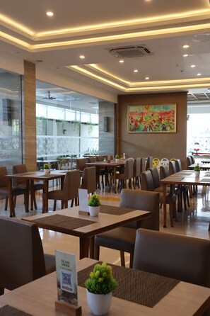 A modern restaurant interior with multiple dining tables and chairs, a large window with plants, and a colorful painting on the wall.