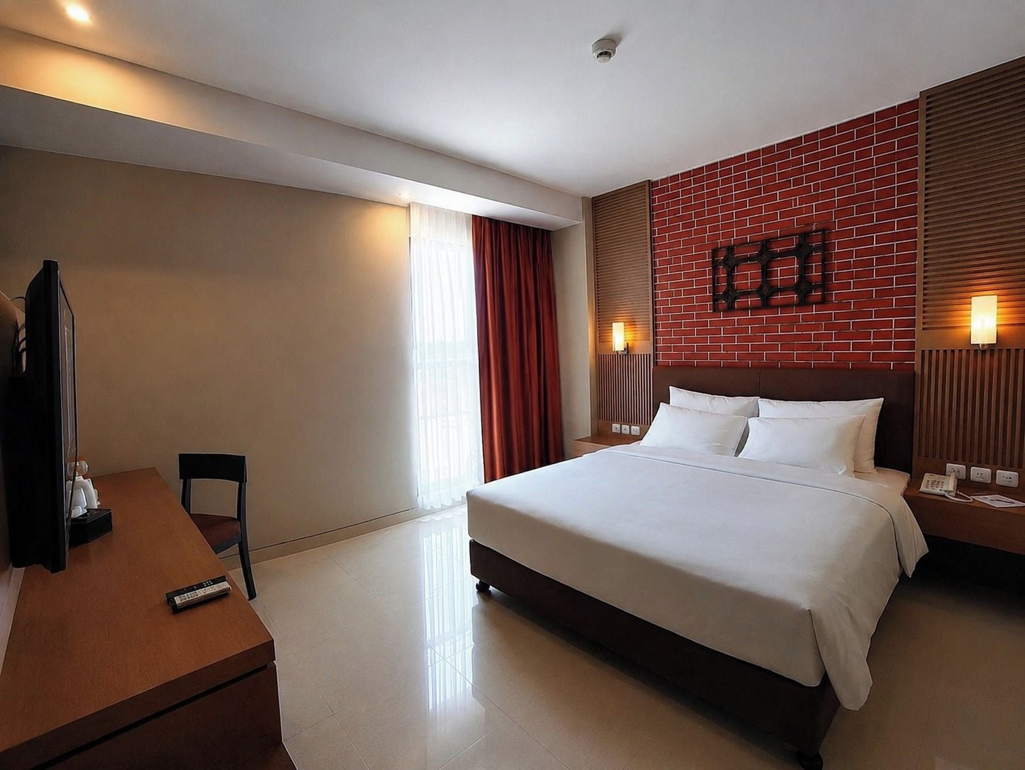 A hotel room with a bed, a desk, a chair, a TV, and a red brick accent wall.
