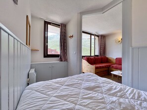 A bedroom with a bed, a window with curtains, a wooden bench with a red cushion, and a small table with a glass. - (BO.130) Family-friendly two-bedroom apartment with a view of the slopes, Les Menuires (Les Menuires)