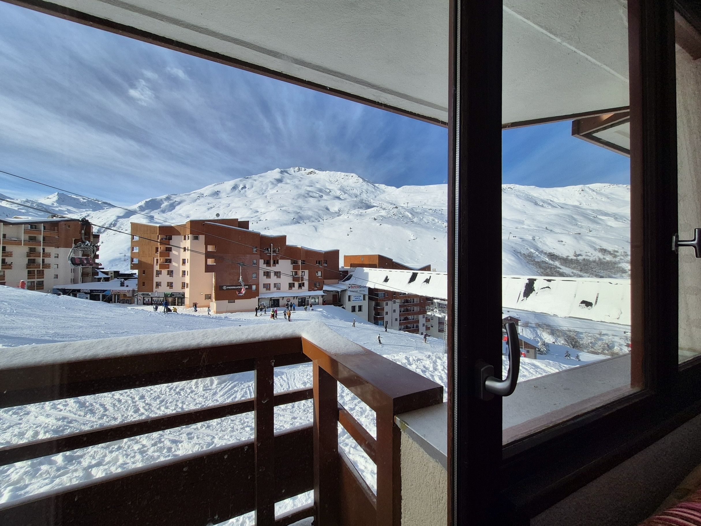A snowy mountain view from a balcony with a wooden railing, a ski lift in the background, and buildings with signage.