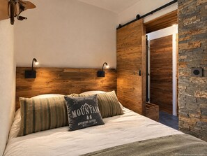A bedroom with a wooden headboard, two wall-mounted lights, a bed with a mountain-themed pillow, and a sliding door leading to another room. - (CI.17B) – Alpine Lodge - Premium "Skis on" Package (Les Menuires)