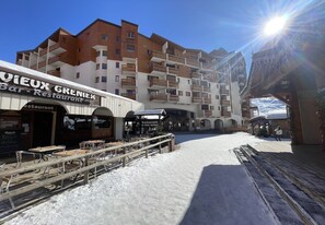 A snow-covered outdoor seating area with tables and chairs, a building with balconies, and a sign for 'Vieux Grenier Bar-Restaurant'. - (CI.17B) – Alpine Lodge - Premium "Skis on" Package (Les Menuires)