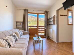 Ein Wohnzimmer mit einer Couch, einem kleinen Tisch mit Buch und Gläsern, einem an der Wand montierten Fernseher und einem Fenster mit Vorhängen.
