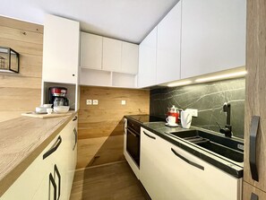 Cuina petita amb armaris blancs, taule negra i un fons de vidre de fusta.