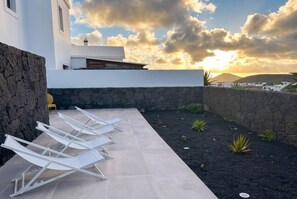 Una terrassa al terrat amb seients de relaxació blancs, una paret de pedra i vista al mar al capvespre.
