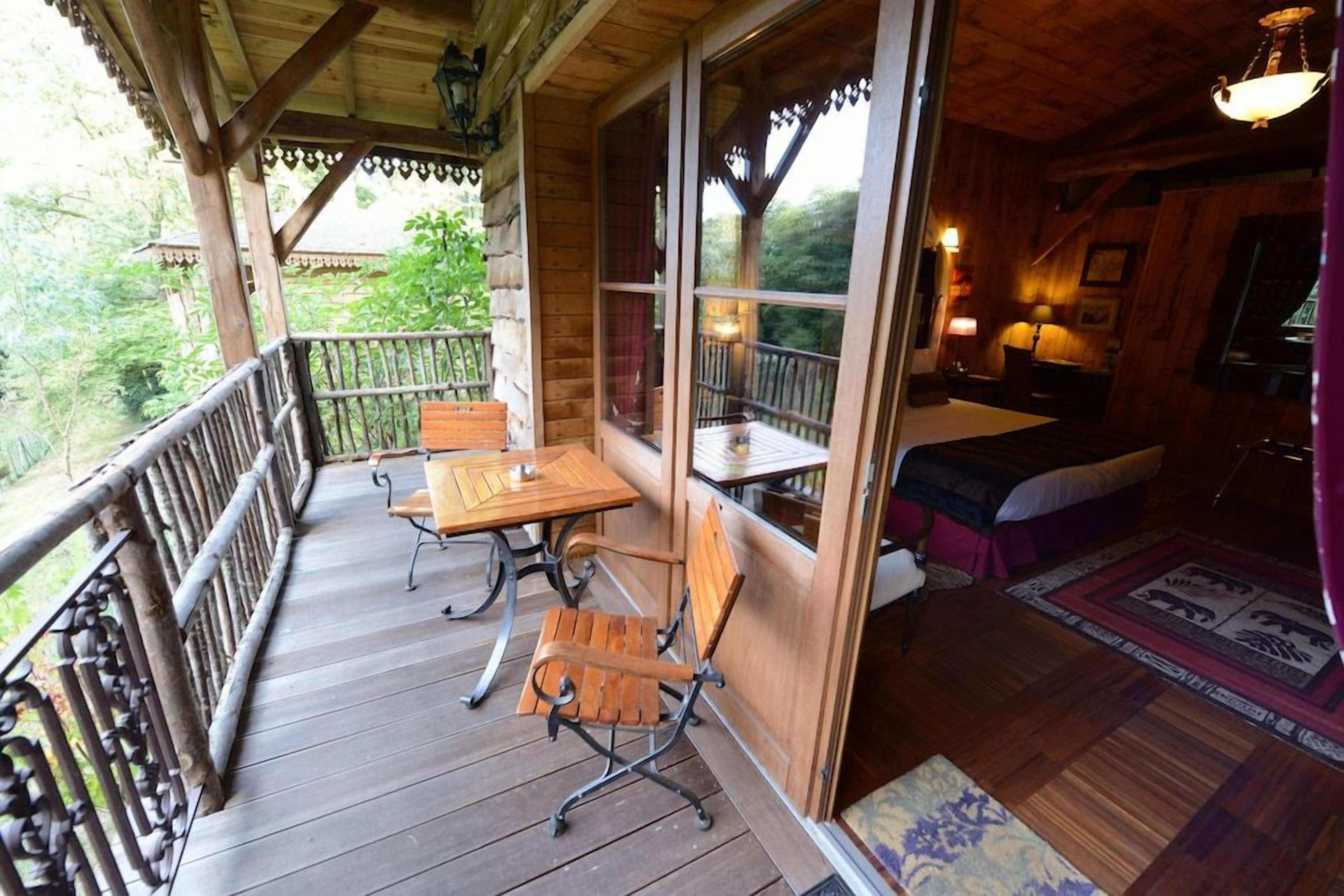 A wooden deck with a table and chairs, a small room with a bed and a window, and a view of greenery outside.