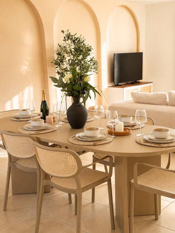 Un coin repas avec une table ronde dressée pour quatre personnes, entourée de chaises en osier. Un vase avec des plantes vertes est posé sur la table, et une bouteille de champagne est visible.