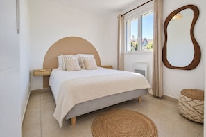 Une chambre à coucher avec un lit, une fenêtre avec des rideaux, un miroir et un tapis rond en fibres tressées.