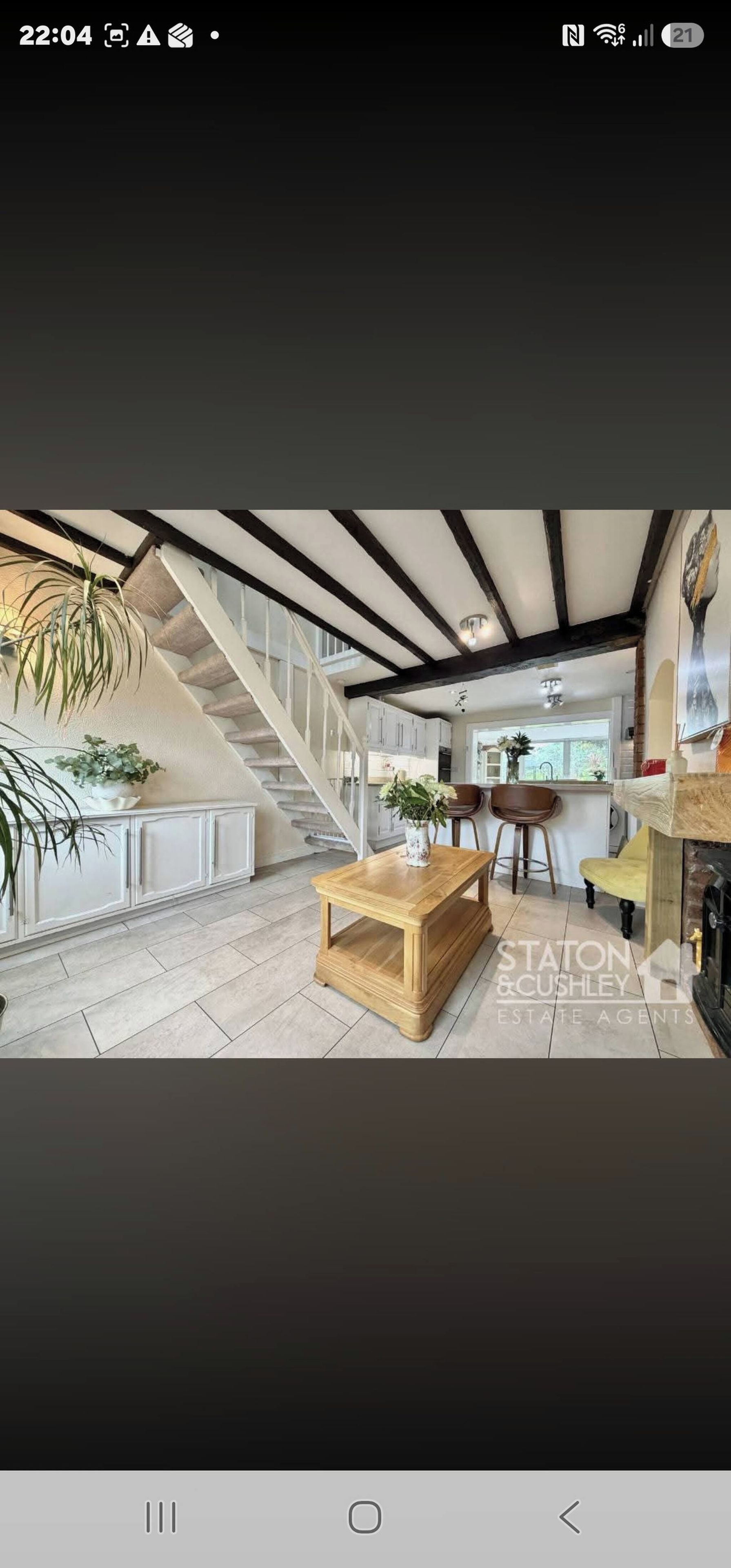 Un escalier menant à une cuisine avec des meubles blancs, une table en bois ornée d’un vase et deux tabourets de bar.