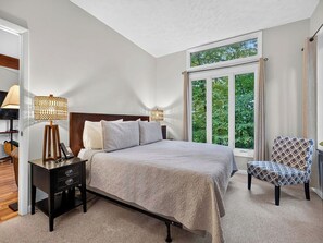 Ein Schlafzimmer mit einem großen Bett, Nachttischlampen, einem Sessel und Blick auf Grün durch ein Fenster.