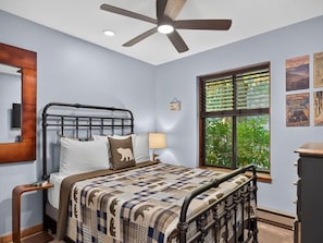 Une chambre à coucher comprenant un lit, une table de chevet, un ventilateur de plafond, une fenêtre avec des stores et des éléments de décoration murale.
