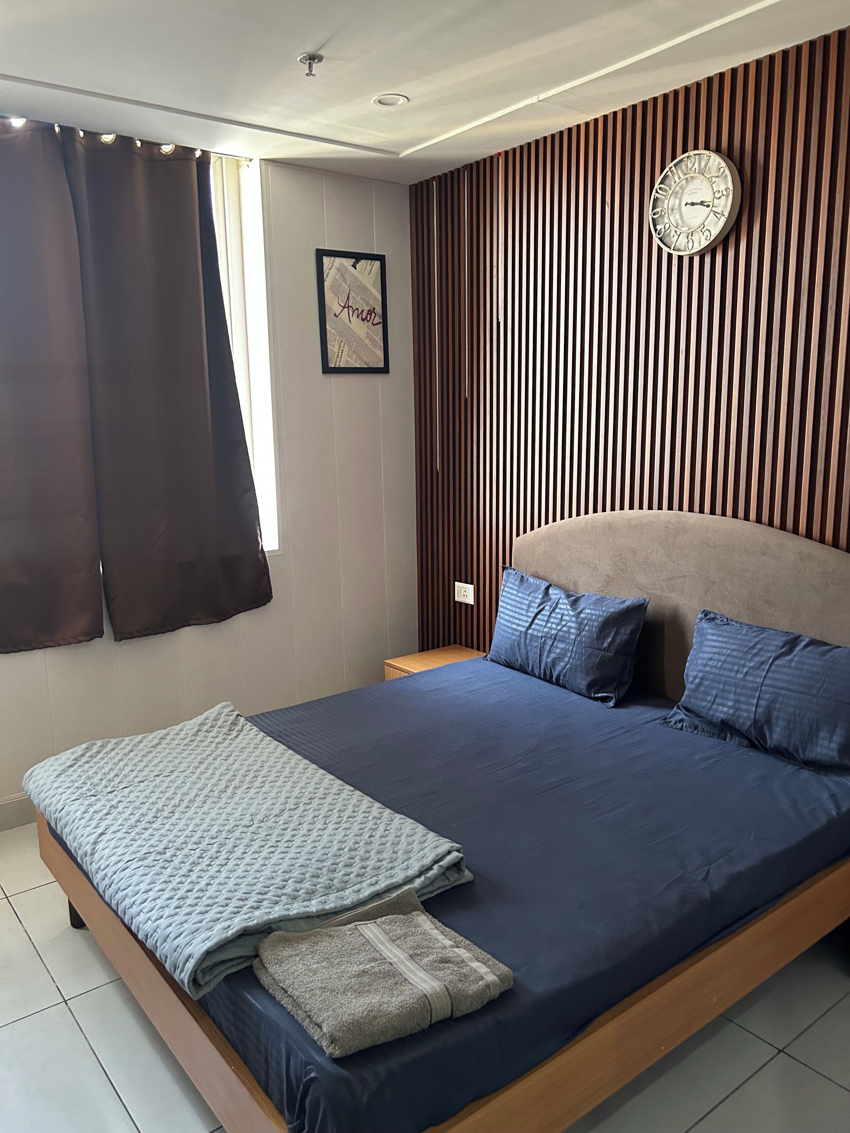 Ein Schlafzimmer mit einem großen Bett, einer Wanduhr, einem gerahmten Bild und einem Fenster mit Vorhängen.