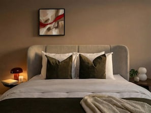 Un lit bien fait, avec une tête de lit grise, deux oreillers et une couverture. On aperçoit une table de chevet avec une lampe et un livre.