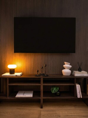 Un salon moderne avec un meuble TV en bois foncé, une lampe de table allumée et un téléviseur à écran plat fixé au mur.
