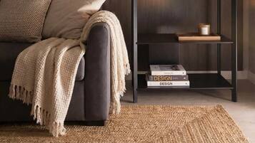 Ein modernes Wohnzimmer mit dunkler Holzvertäfelung, einem dunklen Sofa mit beiger Decke, einem gewebten Teppich und einer schwarzen Regaleinheit mit Dekoration und Büchern.