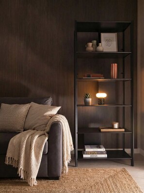 Un salon moderne avec un mur recouvert de panneaux en bois foncé, un canapé sombre agrémenté d’un plaid beige, un tapis tissé et une étagère noire où sont disposés des objets décoratifs et des livres.
