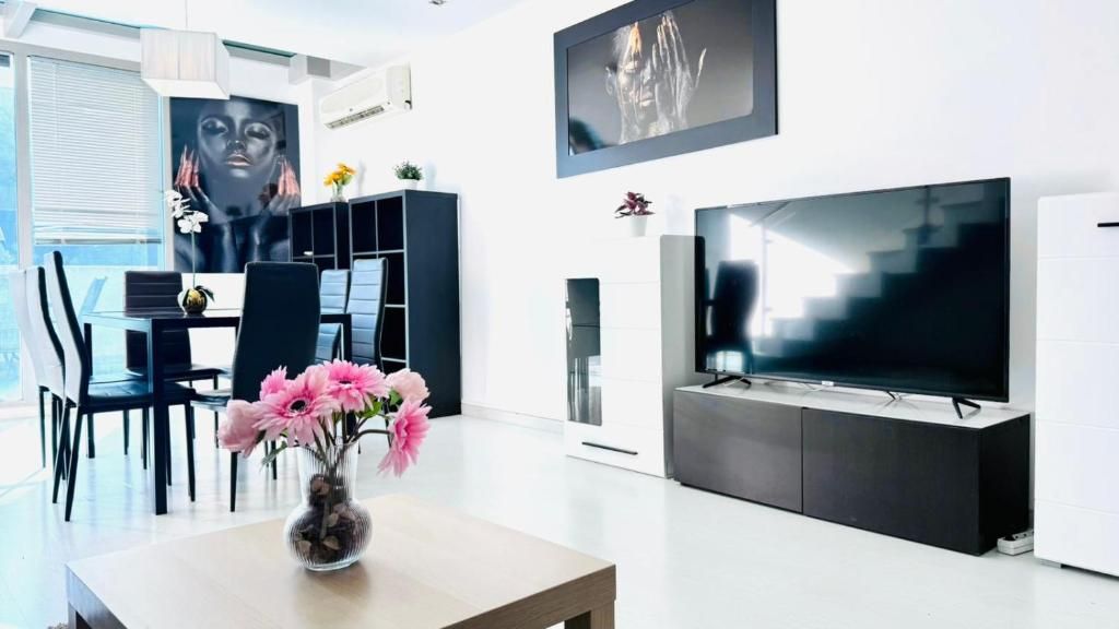 Un salon moderne doté d’un téléviseur à écran plat, d’un coin repas avec des chaises, d’un vase de fleurs et d’une grande fenêtre.