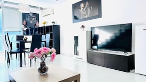 A modern living room with a flat-screen TV, a dining area with chairs, a vase of flowers, and a large window. - Lumen Triplex (Madrid)
