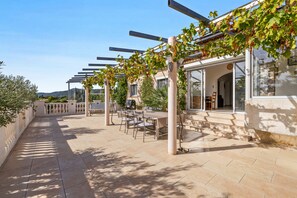 Et overdækket udendørs opholdsområde med borde og stole, en pergola med slyngplanter og udsigt over det omkringliggende landskab.
