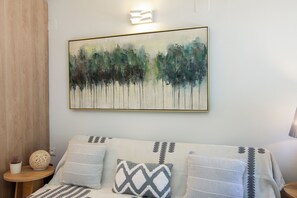 Ein Zimmer mit einem Bett, Kissen, einem kleinen Tisch mit Pflanze und einem Bild an der Wand.