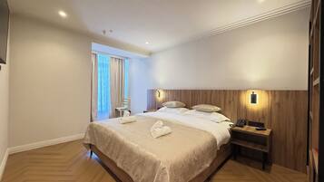 Habitación de hotel con una cama grande, cabecera de madera, mesitas de noche, una silla y una ventana con cortinas.
