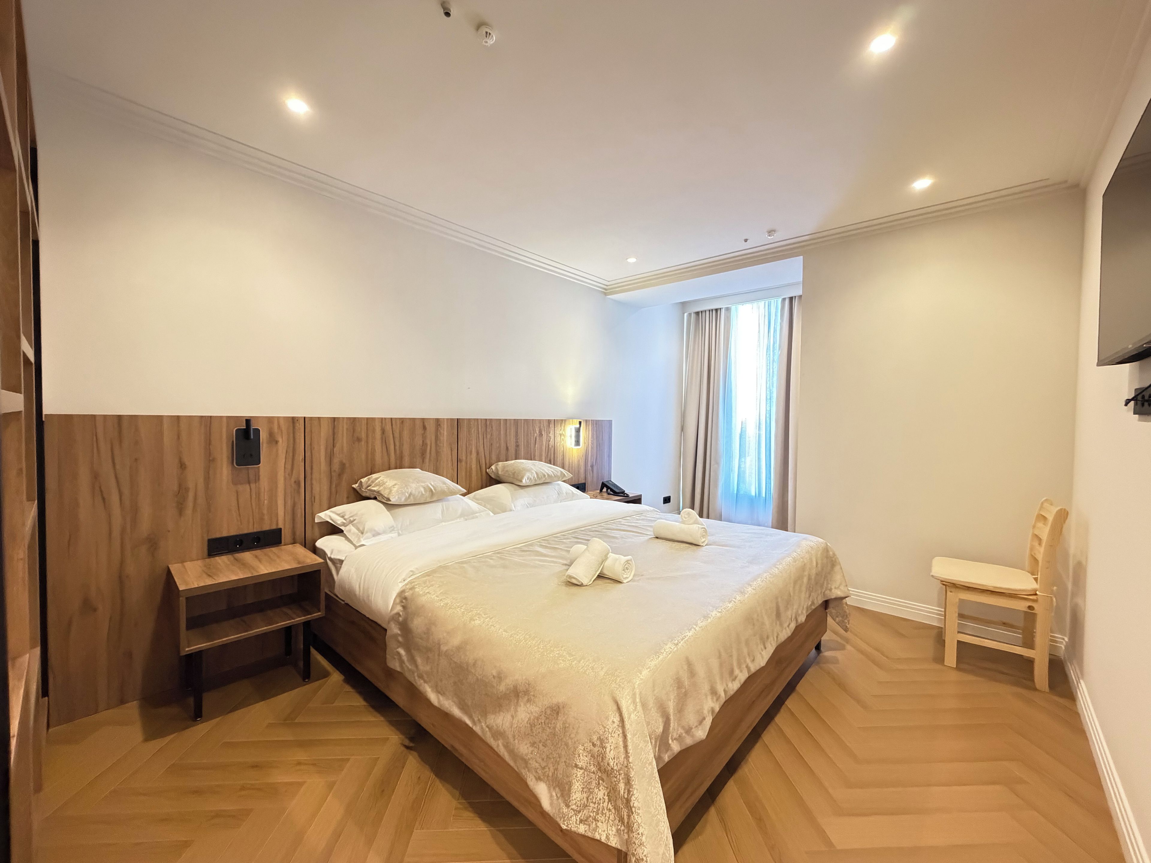 Habitación de hotel con una cama grande, cabecera de madera, mesita de noche, una silla y televisor de pantalla plana montado en la pared.