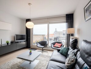 A modern living room with a sofa, a coffee table, a TV, and a balcony with a view of buildings. - Nice apartment for 2 people with WIFI, TV and terrace (Blankenberge)