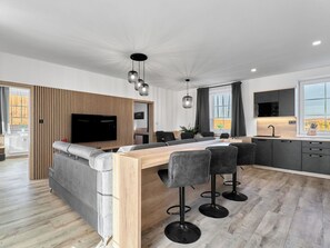 A modern kitchen with a central island, bar stools, a dining area, and a built-in TV.