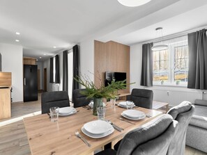 A modern dining area with a wooden table set for four, a television, and a view of a field through large windows. - Beautiful apartment for 5 people with WIFI, TV, patio and panoramic view (Klášterec Nad Ohří)