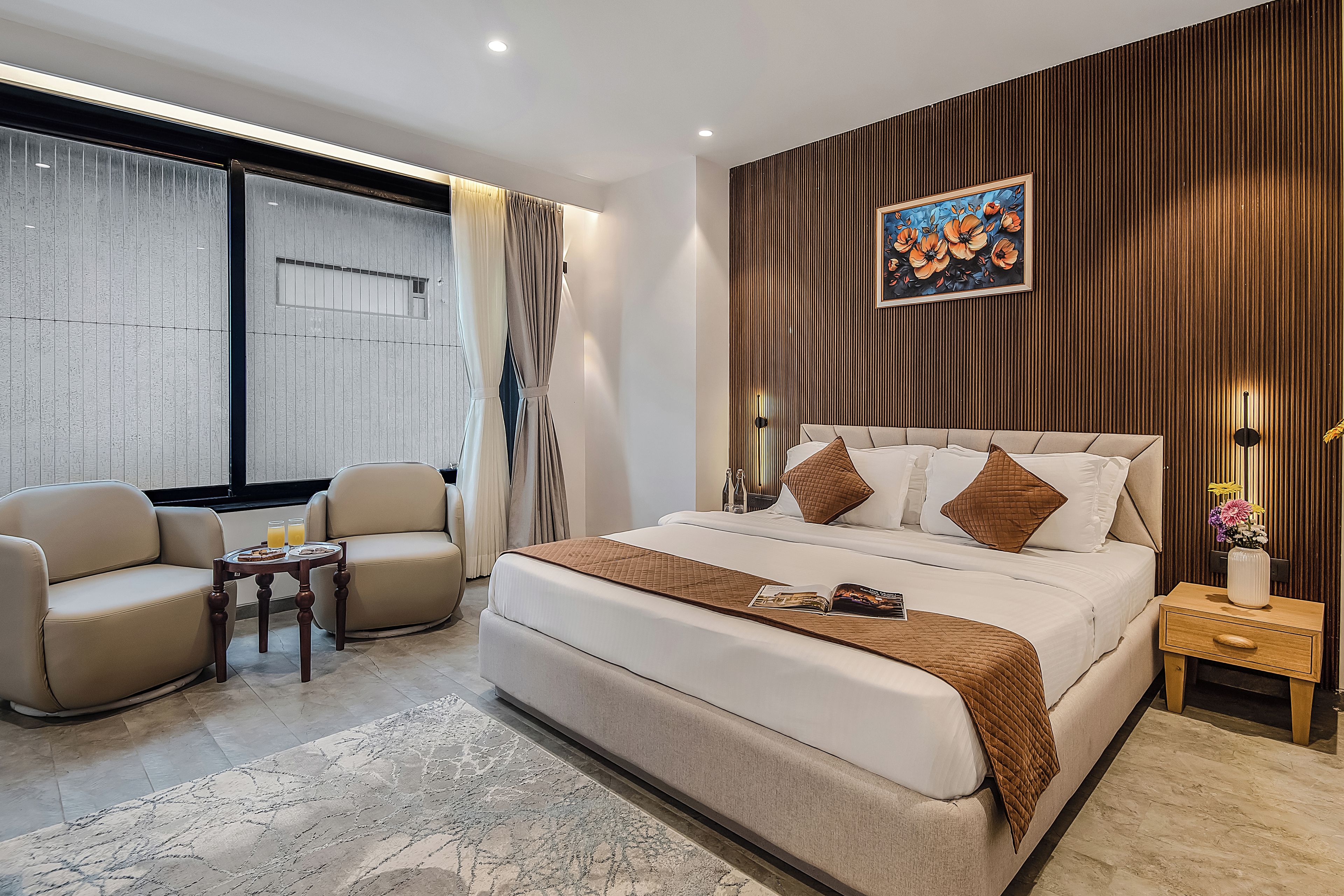 Un dormitorio moderno con una cama grande, dos sillones, una mesita con bebidas y un panel decorativo en la pared.