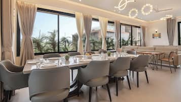 A modern dining area with a long table, multiple chairs, and a view of palm trees through large windows.