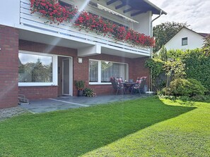 A two-story residential building with a balcony adorned with red flowers, a patio with outdoor seating, and a well-maintained lawn. - Lovely apartment for 3 people with WIFI, TV and terrace (Raben Steinfeld)