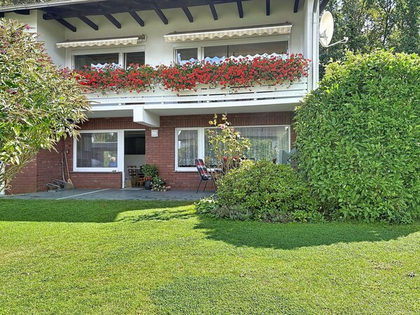 A two-story building with a balcony adorned with red flowers, a brick facade, and a well-maintained lawn. - Lovely apartment for 3 people with WIFI, TV and terrace (Raben Steinfeld)