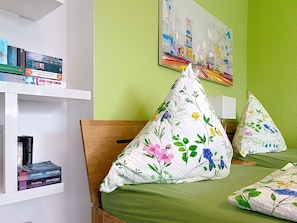 Een kamer met een bed, kussens, een boekenkast en een schilderij aan de muur.