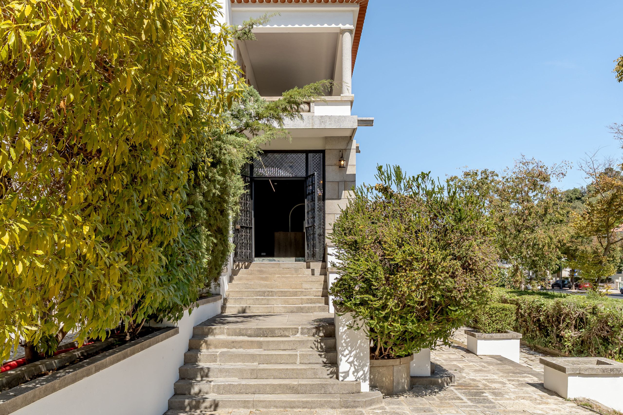 A building with a staircase leading to a dark entrance, surrounded by greenery and a clear sky.