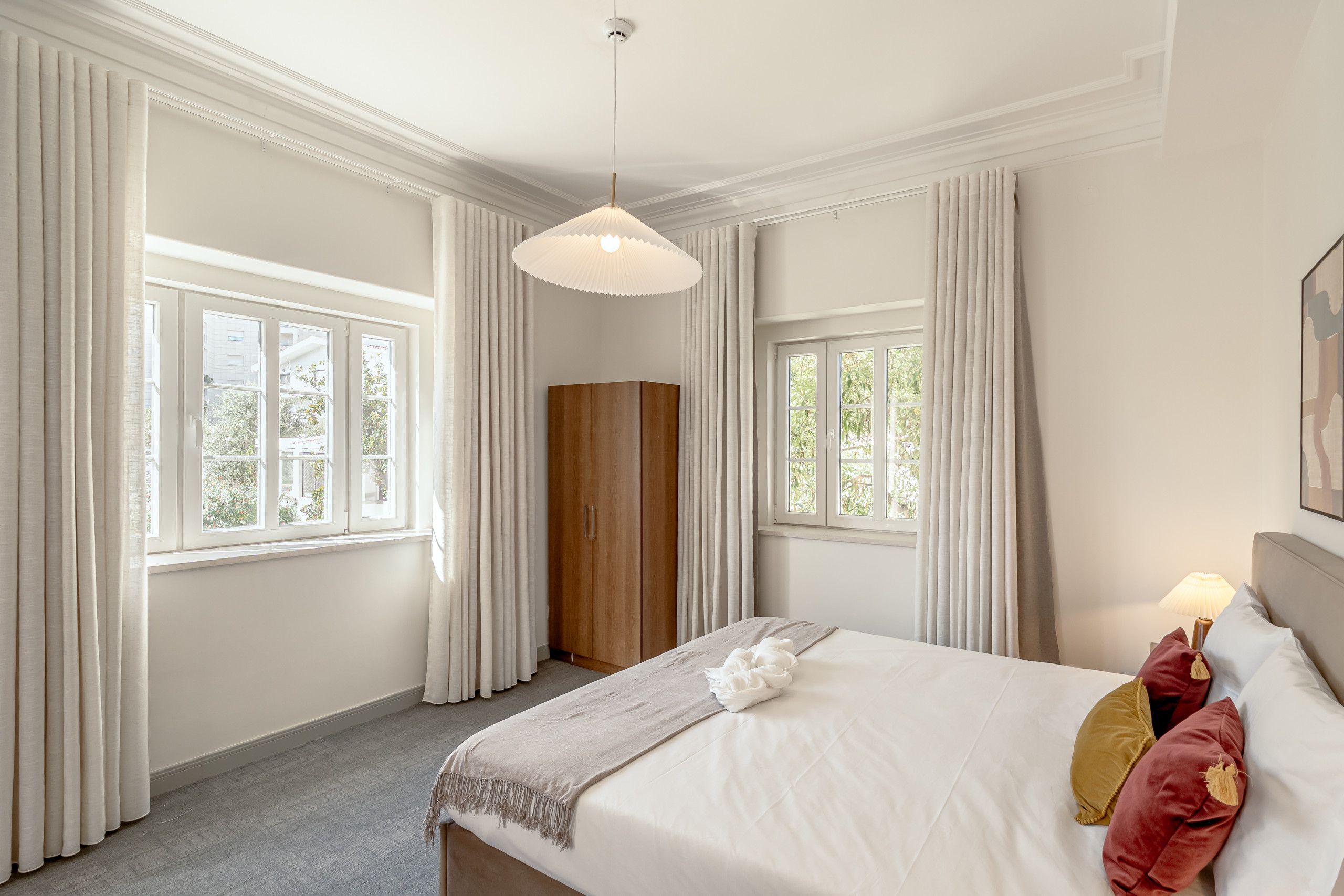 A hotel room with a bed, a wardrobe, and two windows with curtains.