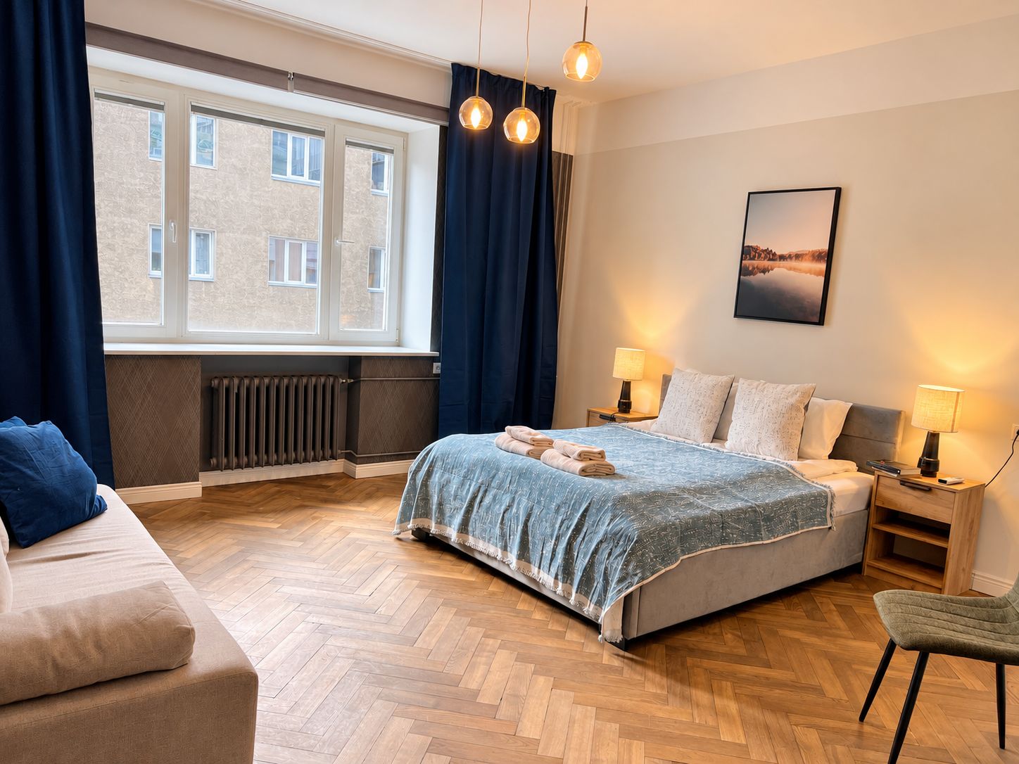 Ein modernes Hotelzimmer mit einem großen Bett, zwei Nachttischen mit Lampen, einem Sofa, einem Sessel und Blick auf ein Gebäude durch ein Fenster.