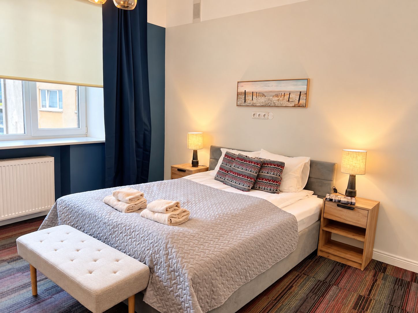 Quarto de hotel com cama, duas mesinhas de cabeceira, um banco, janela com cortinas e um quadro na parede.