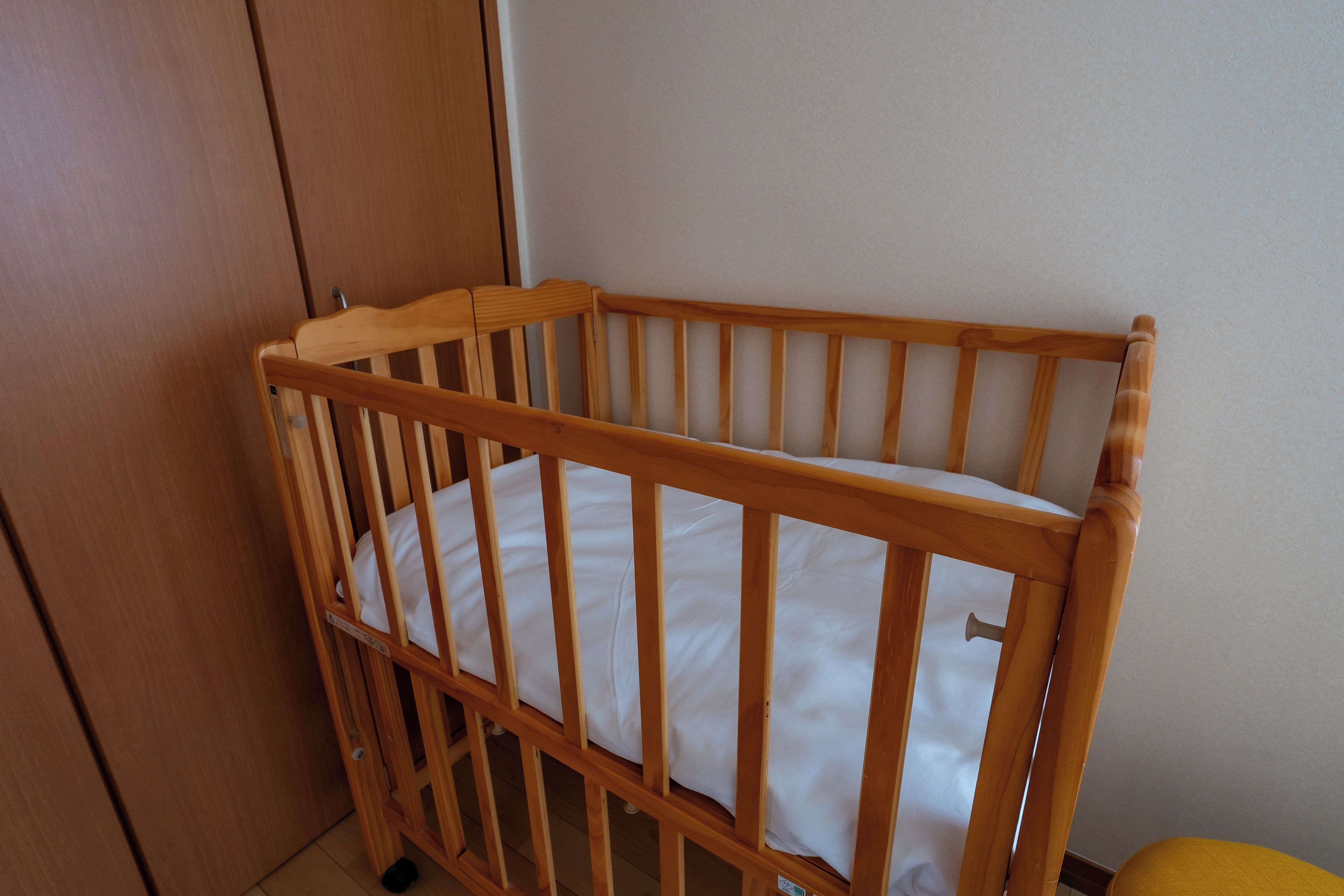 Un berceau en bois avec un matelas blanc, placé contre un mur où se trouve une porte, et un petit objet jaune sur le sol.