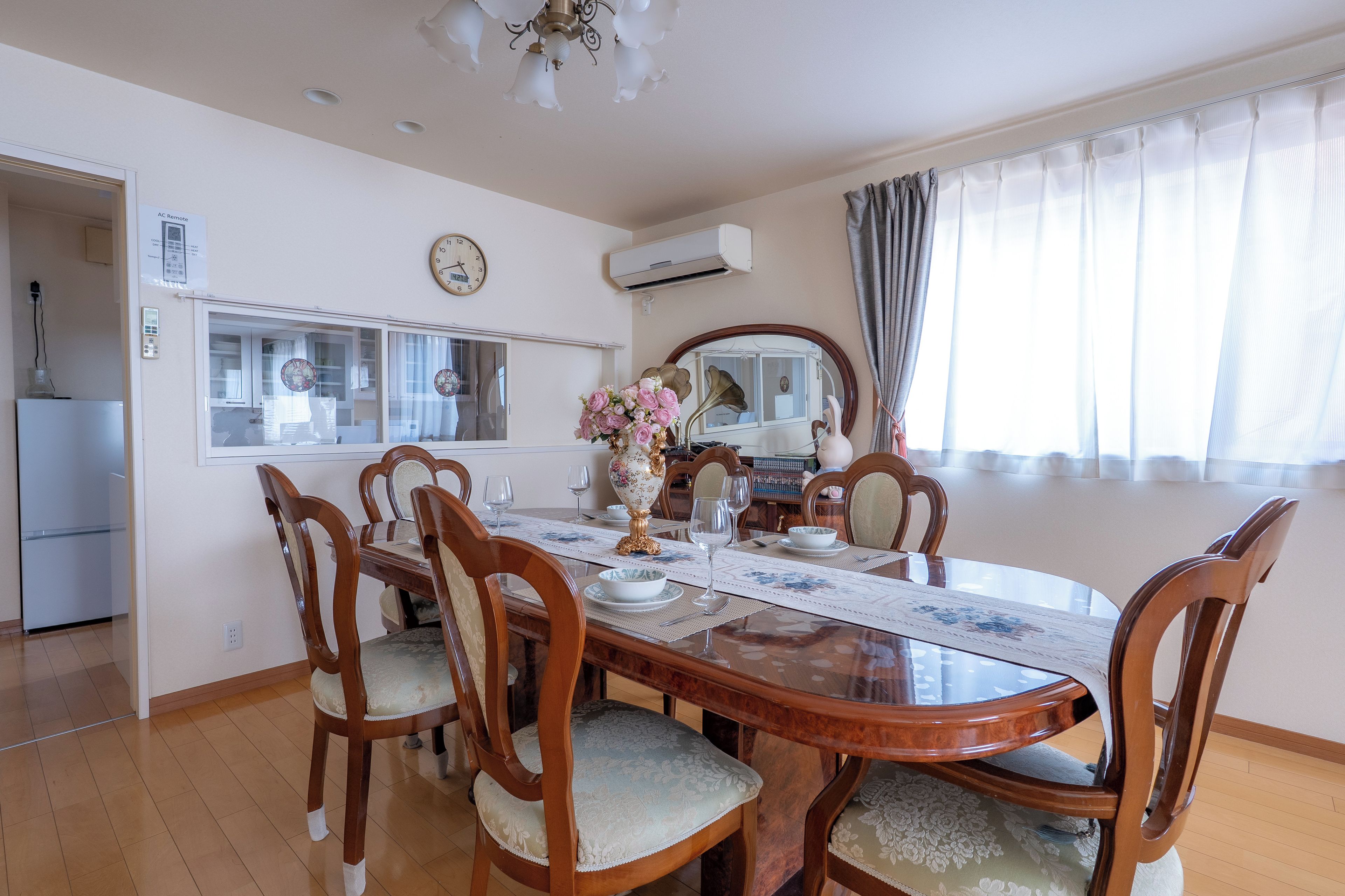 A dining area with a wooden table, chairs, a clock, and a window with curtains.