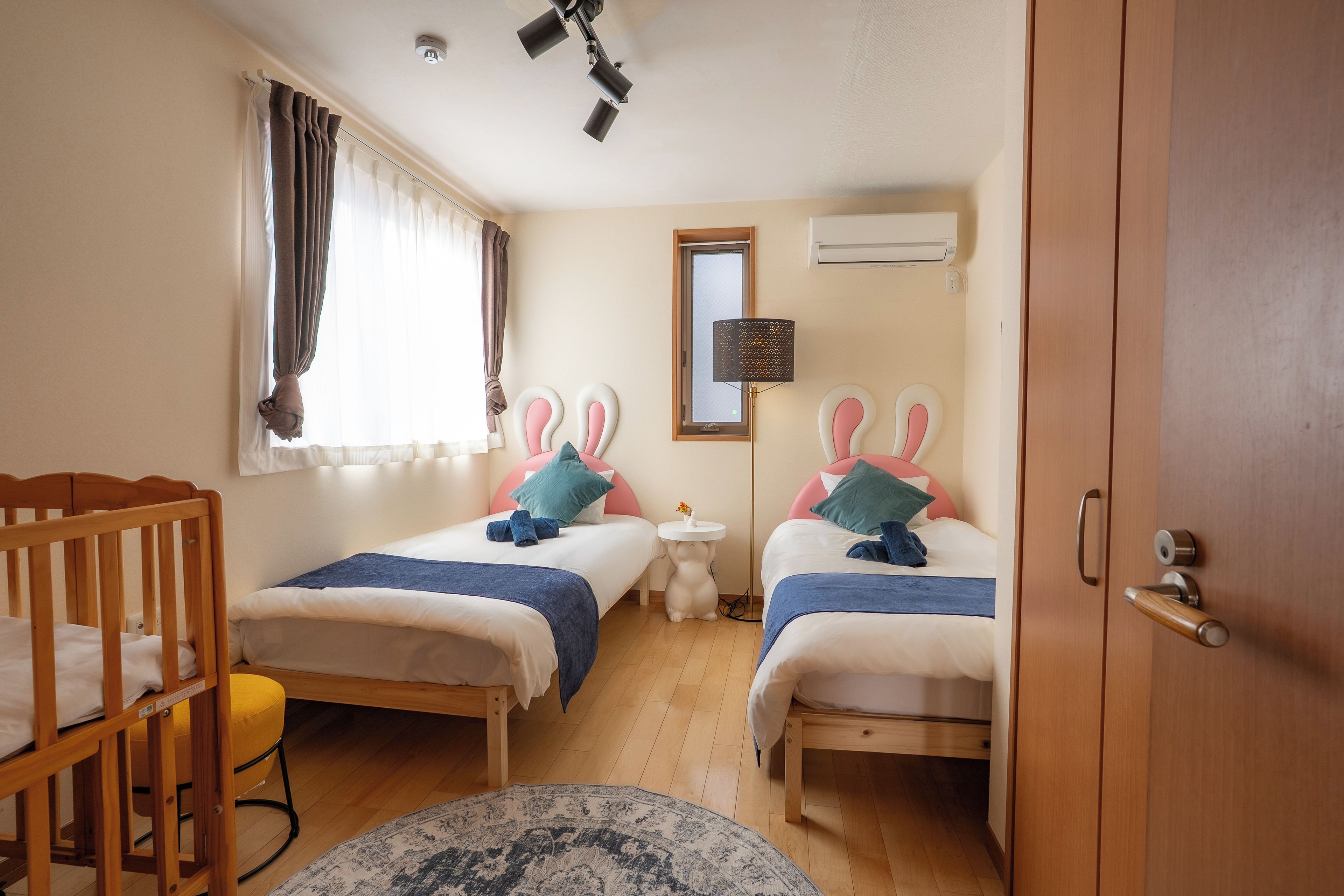 A bedroom with two beds, a crib, a window with curtains, a bedside table, a mirror, and an air conditioning unit.