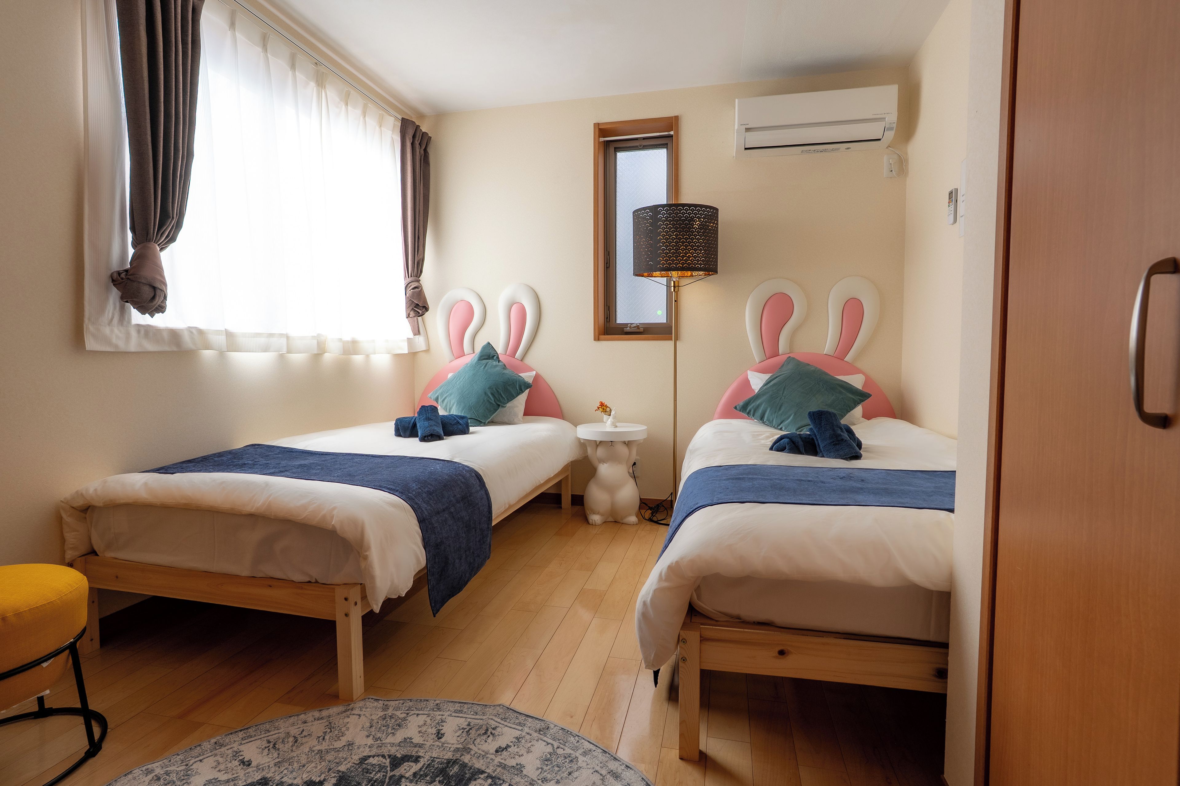 A hotel room with two beds, a window with curtains, an air conditioning unit, and a small round table.