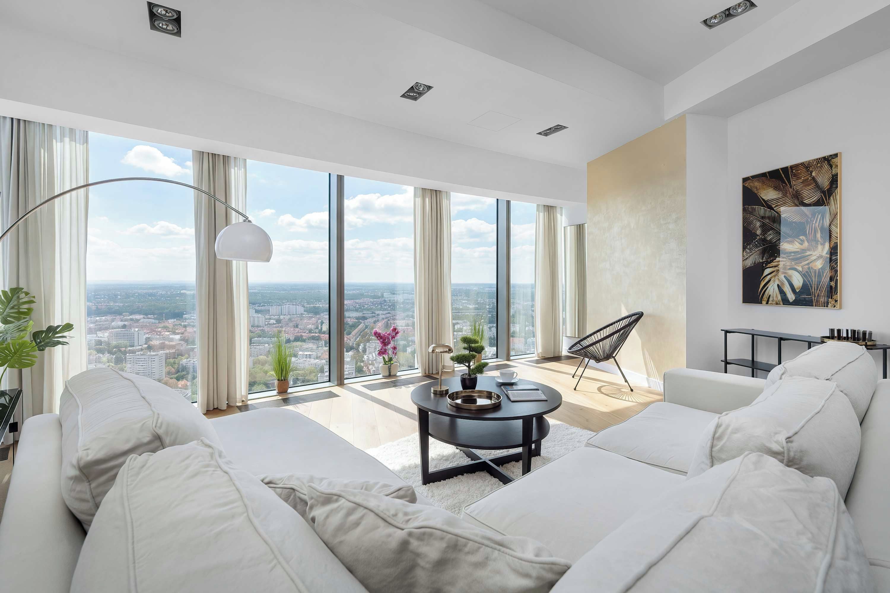 A modern living room with a large window, white furniture, a coffee table, and a view of a cityscape.