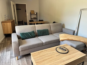 Ein Wohnzimmer mit einem grauen Ecksofa, zwei grünen Kissen, einem Holztisch mit Fernbedienung und einem weiteren Holztisch.