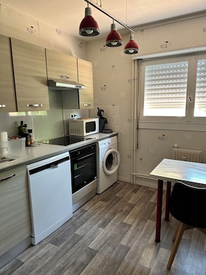 Eine kompakte Küche mit Geschirrspüler, Mikrowelle und Waschmaschine. Ein Fenster mit Jalousien, ein Tisch und ein Stuhl sind vorhanden.